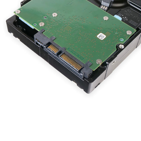 A close-up, angled view of the underside of a 3.5-inch internal hard drive. A green printed circuit board (PCB) is visible, showing a Serial ATA (SATA) interface with gold-plated data and power connector pins. The drive's main body is made of a solid black material, and a thin, dark ribbon cable connects the circuit board to the internal spindle motor. Small technical markings and a QR code are visible on the green board.